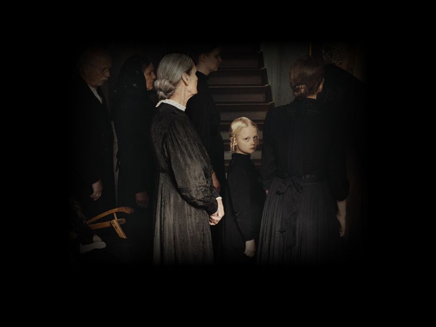 A solemn Victorian child in a black dress stands amongst women in black dresses