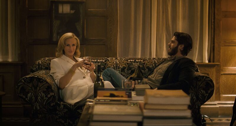 A man and a woman sit on a couch in front of a pile of books, drinking wine.