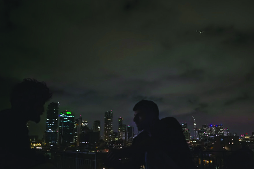 Two people on a rooftop in a city at night. There are lights in the sky.