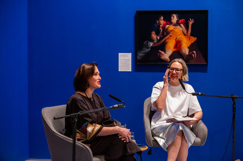 Two white women sit in chair during a talk with microphones in front of them - a blue wall with a painting in the background 