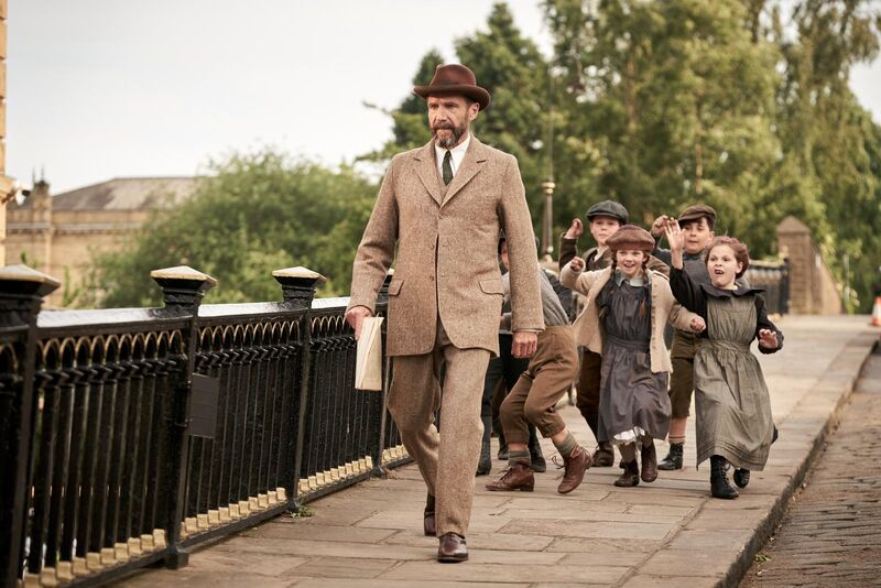 A man in a beige suit walks down a street, while a group of children in 1910s clothing chase him shouting