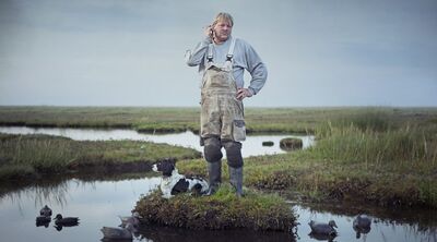 A blonde man in overalls sands on a patch of grass in the middle of a marshy plain with a dog at his feet.