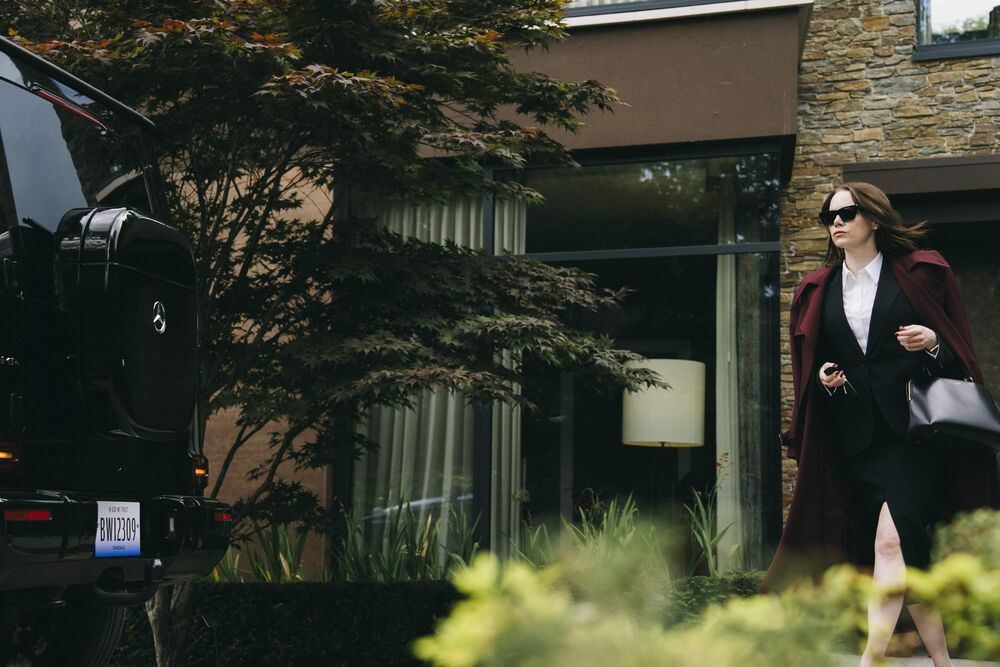 A woman wearing a red jacket and blakc sunglases walks towards a large black car