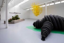 A gallery space with a black sculpture of the Michelin made from tyres lying flat on a green shape, plants and a yellow cloud hanging from the ceiling