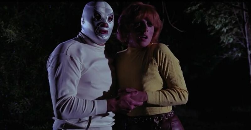 Man and woman holding each other while standing outside in the dark. The man wears a white Lucha libre mask and a white jumper. The woman has red hair and wears a yellow top