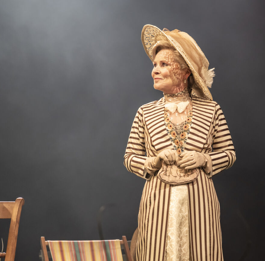 A woman in a large straw hat and a stripy 1900s dress stands on stage.