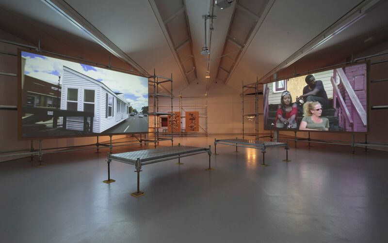 DCA Gallery displays two large scaffold structures with televisions on them, as part of Mike Kelley's exhibition. The left television shows a mobile home, and the right television shows three people sitting on the steps outside of a house.