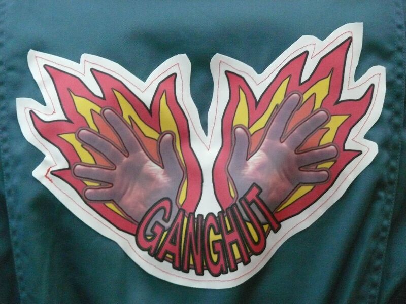 A close-up of the logo on the back of a boiler suit. The logo has two hands with flames behind them, and says 'GANGHUT' in graphic text.