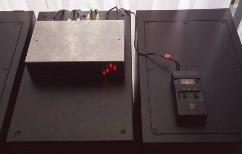 A silver box, with cables plugged into it, sits on a black filing cabinet. One of the cables connects to a small black device, which looks like a tape recorder.