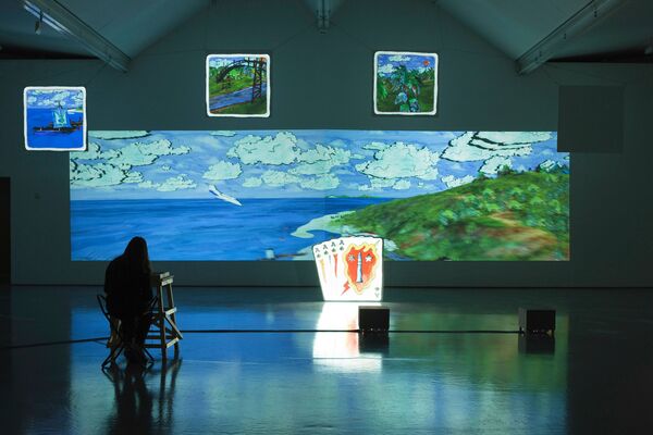 Eddo Stern's exhibition in DCA Galleries. Two large projectors show a painted image of clouds and mountains. An illuminated sculpture of a deck of cards is in front of the projector.
