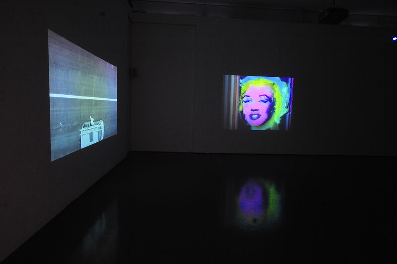 Two screens in DCA galleries from George Barber's exhibition. One screen shows the top of a white building. The other shows a neon-coloured image of Marilyn Monroe. 