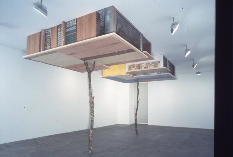 Upside-down, house-like structures hang from DCA Gallery ceiling, propped up by tree branches.