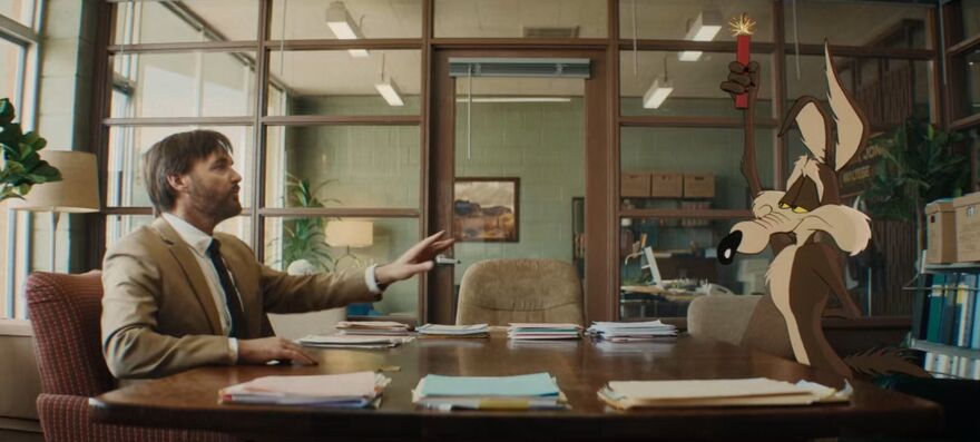A man in a suit talks to a cartoon coyote across a meeting room table