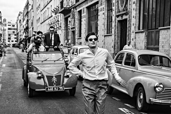 A black-and-white folm stil;l shows three men walking down a Parisian street, with 1960s cars in the background