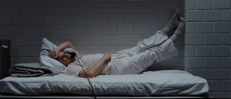 A man in a prison cell lies in bed while talking on the phone