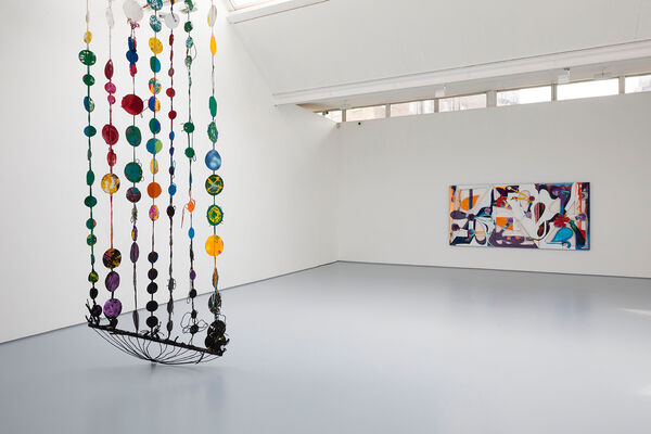 Installation documentation showing a hanging hammock form to the left hand side of the image, a colourful sculpture composed of painted found materials and circle motifs, braided together in strands. In the background to the left of the bright gallery, on a white wall, is a rectangular artwork mainly white, purple, orange and blue. 