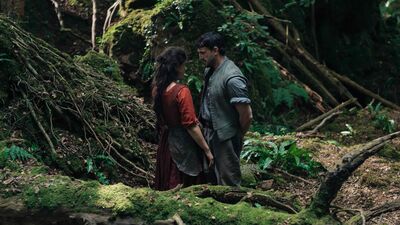 A man and a woman in medieval clothing talk to each other in a forest clearing