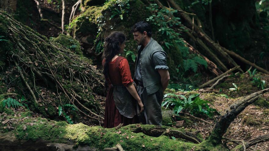 A man and a woman in medieval clothing talk to each other in a forest clearing