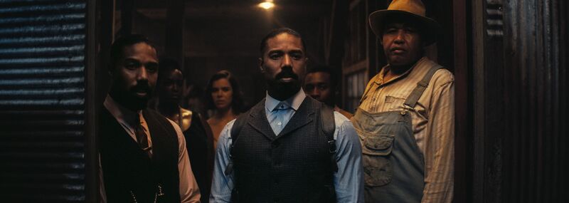 Three men, wearing 1930s style dress, stand at a barn door.