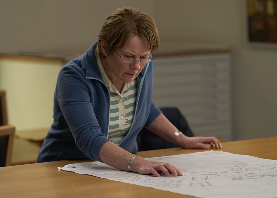 A woman wearing glasses pores over a large document spread out on a desk