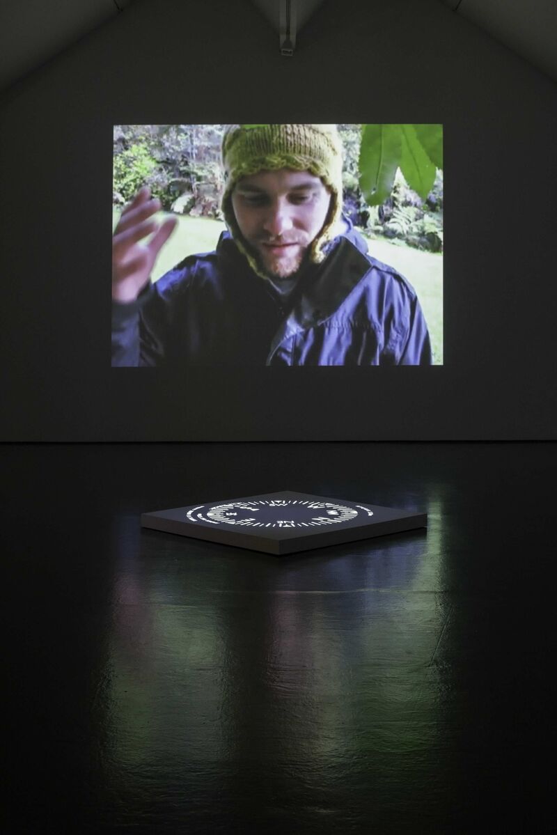 A projector in DCA Galleries from Thomas & Craighead's exhibition shows a man wearing a hat and a raincoat talking.