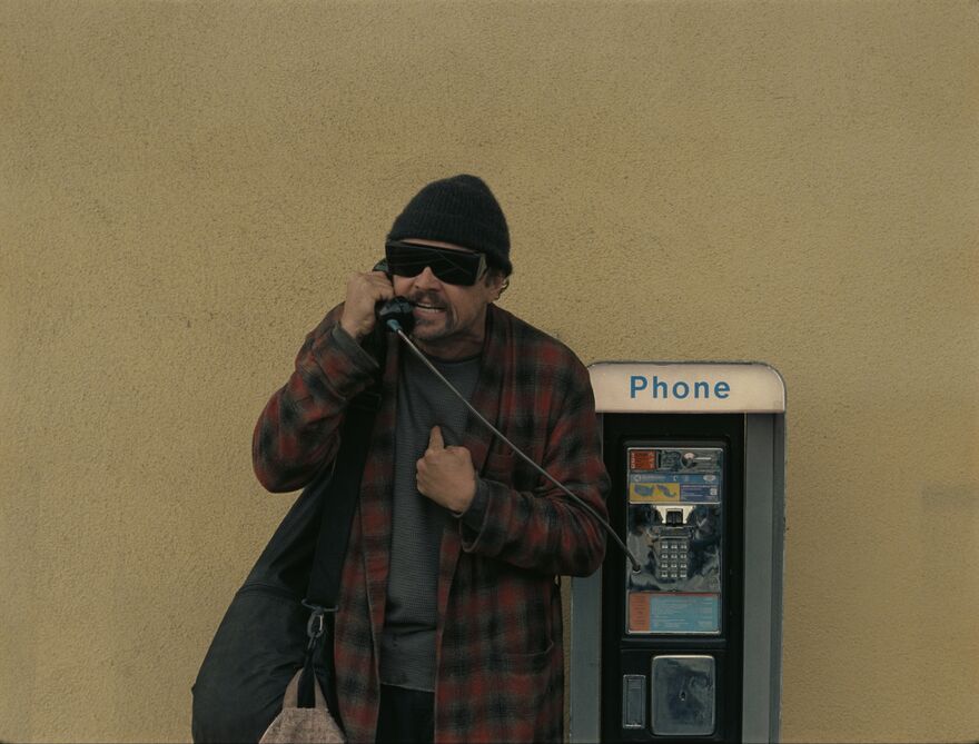 A man wearing a hat and dark glasses looks angry while talking on a public telephone.