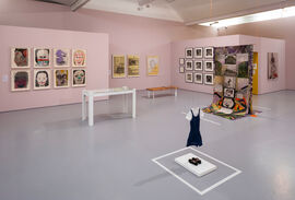 a pink gallery space with paintings prints and photos on the rear wall. In the foreground a small girls uniform and a silk banner 