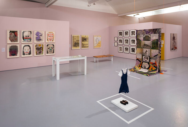 a pink gallery space with paintings prints and photos on the rear wall. In the foreground a small girls uniform and a silk banner 
