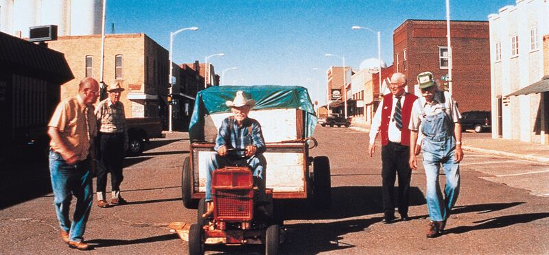 A man rides a lawnmower through a small town, onlookers alongside.