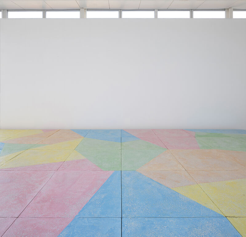 A floor based work is shown in gallery one. The walls are white and the gallery floor is painted grey. On top of this, a sculptural floor is lain in blocks, raising the floor over most of the gallery. This work is brightly coloured, with pastel blue green, yellow orange and pink, in abstract shapes.