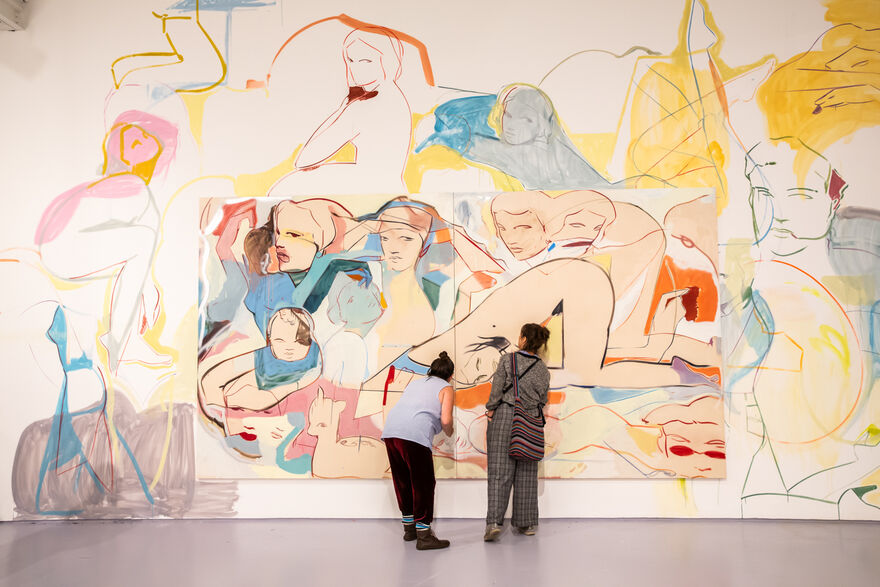 Two figures look closely at a painting that is on a gallery wall with lots of figures painted on ti