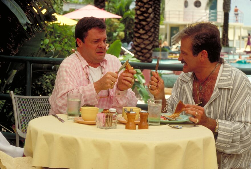 Two men are sat at a table in a tropical setting, discussing cutlery