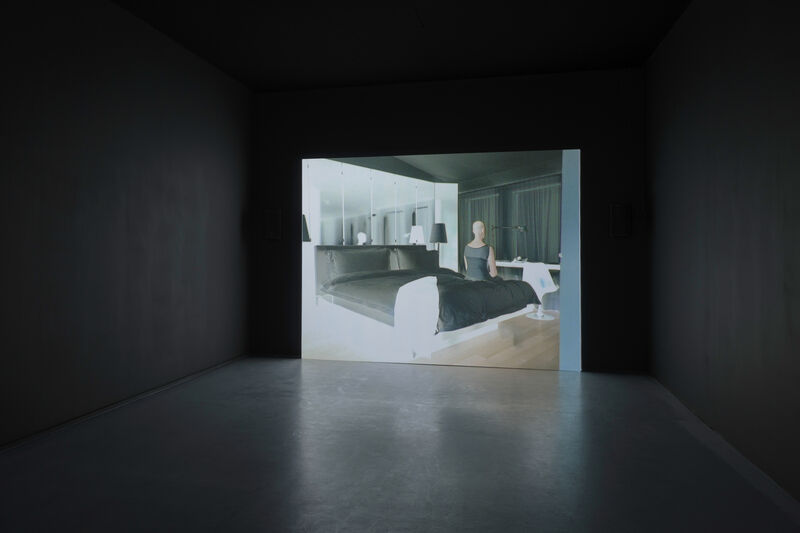 A photograph of a video installation in a small blacked out room. The video shows a figure sitting on a bed, as though in a hotel. The colours are polarised. 