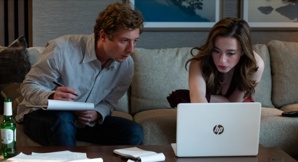 A young man and a young woman sit on a sofa, he's making notes while she's showing him something on her computer
