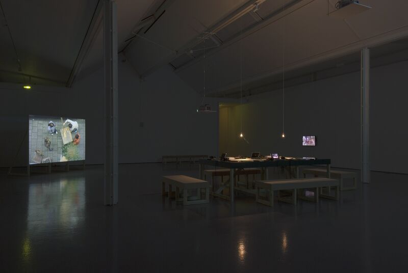 DCA Galleries during Johanna Billing's exhibition. Benches are arranged around a rectangular table. In the distance, a projector shows an image of people working in a garden.
