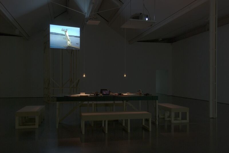 DCA Galleries during Johanna Billing's exhibition. Benches are arranged around a rectangular table. In the distance, a projector shows an image of a crane and a body of water.