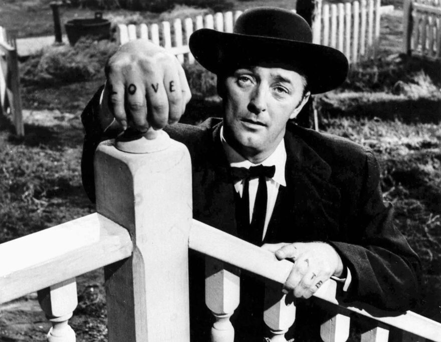 A man wearing a suit and hat, with the word 'LOVE' tattooed on his fingers, leans on a bannister.