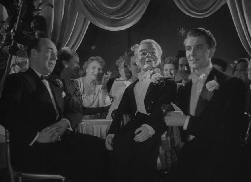 A man with a ventriloquist dummy perfroms in front of tables of people