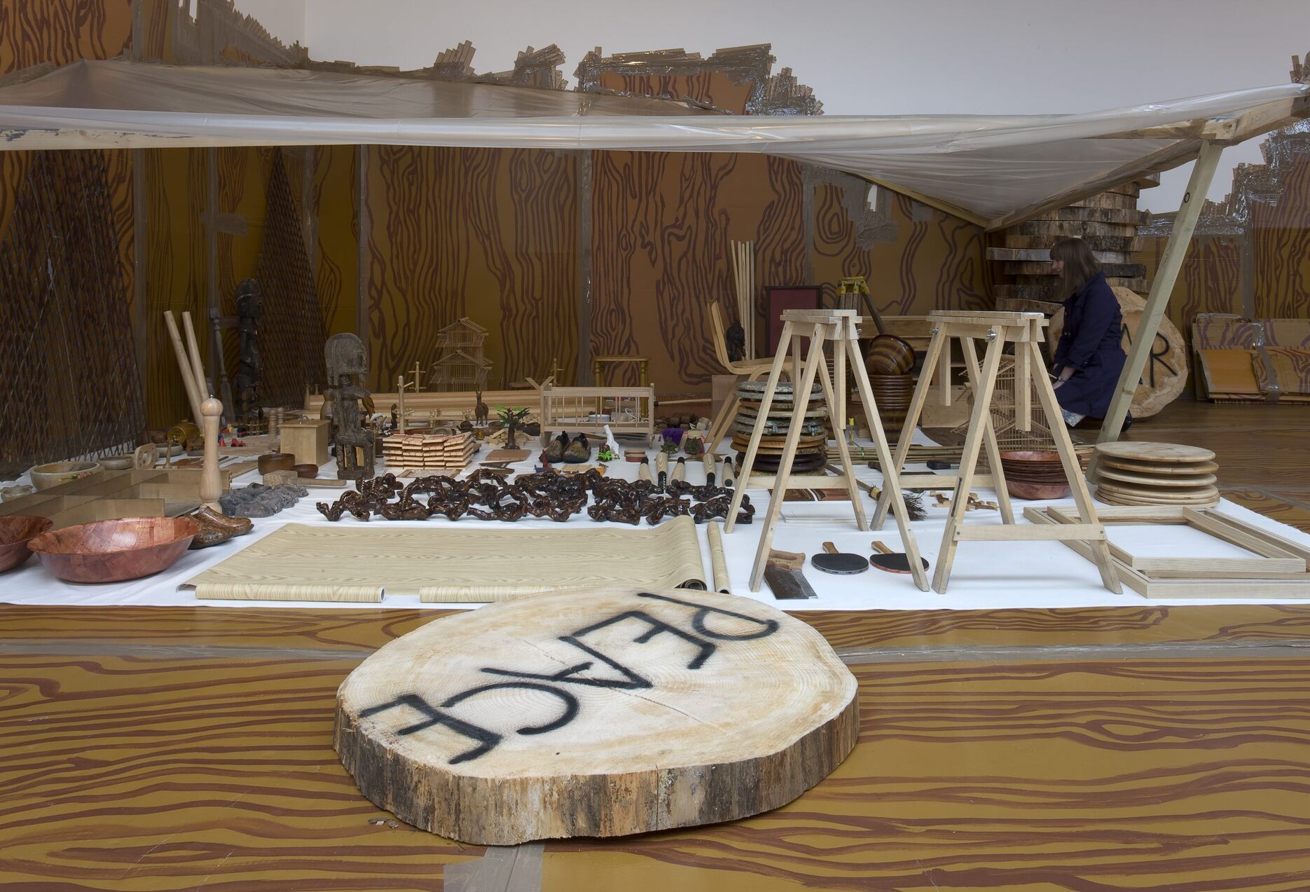 From Thomas Hirschhorn's exhibition. There is a collection of small wooden ornaments and baskets on a mat on the floor. There's also a tree stump with the word 'PEACE' spray painted on it.