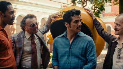 A man stands next to a group of other men who are all staring at him. Everyone is wearing 1970s clothes