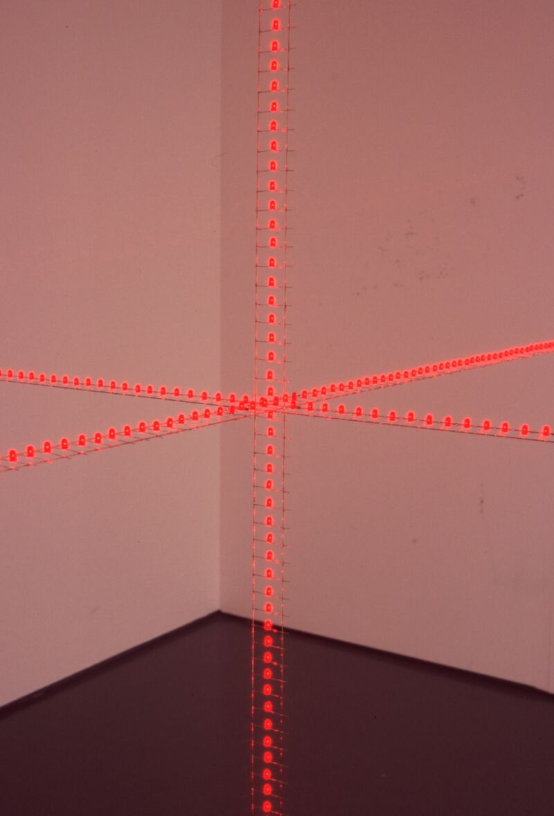 Work by Grönlund\Nisunen - orange thread is arranged in a cross shape in front of a wall.