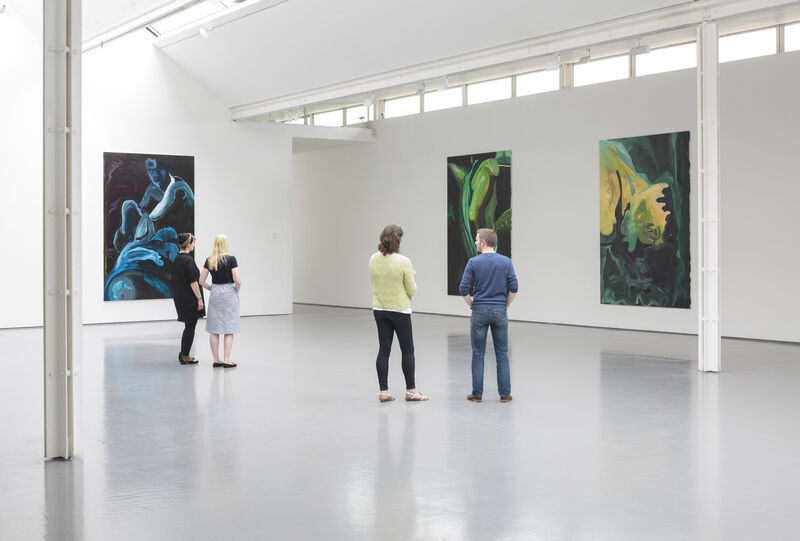 Installation view of a gallery with bright natural light and white walls. We can see three paintings from a distance. The paint strokes are bold and broad. A few people look at the paintings, dispersed through the gallery.