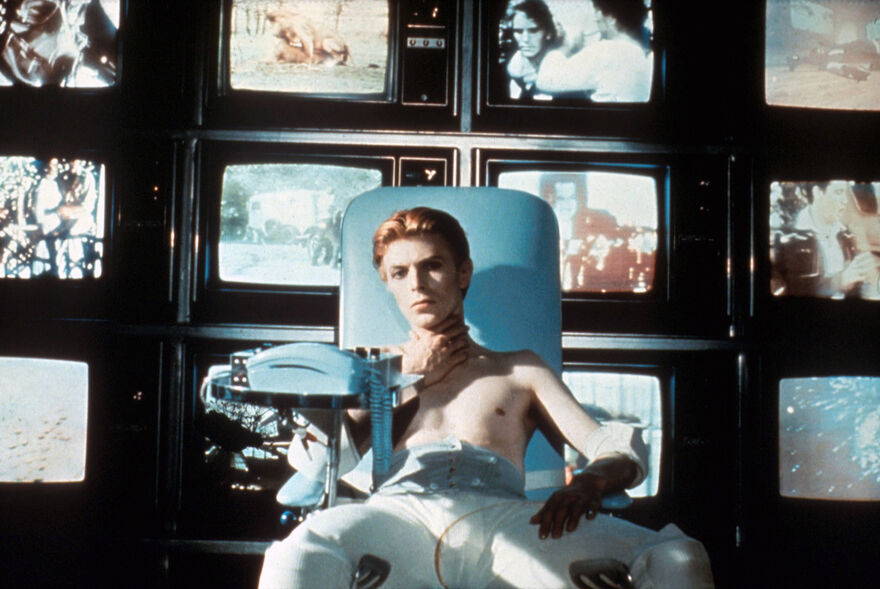 A shirtless man sits in front of a bank of TV screens