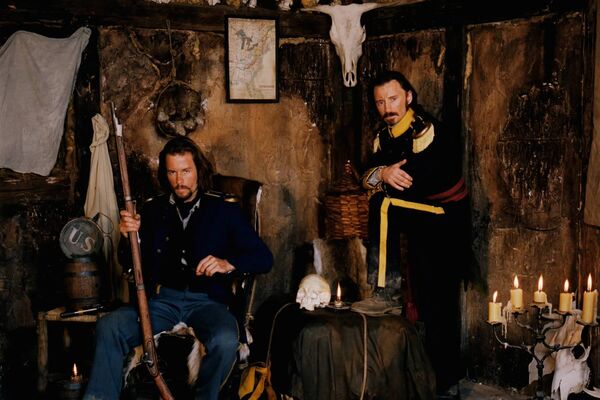 Two men in 19th century American clothes stand in an abandoned cottage.