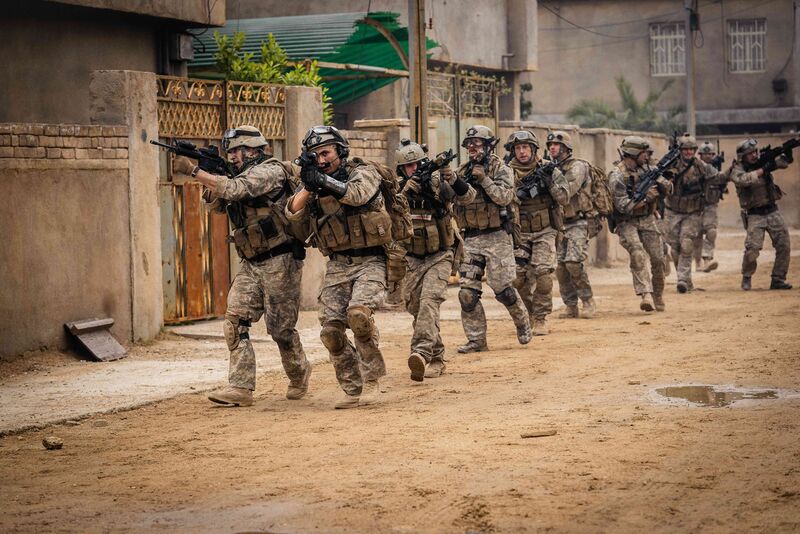 A group of soldiers in camouflage, holding guns, walk around a sandy neighbourhood.