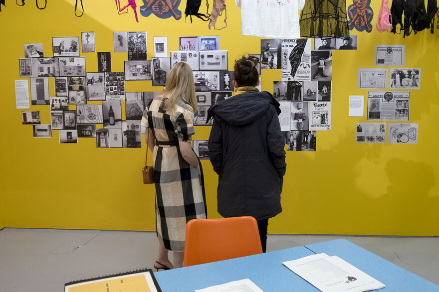 A yellow wall with pictures on it being viewed by two people 