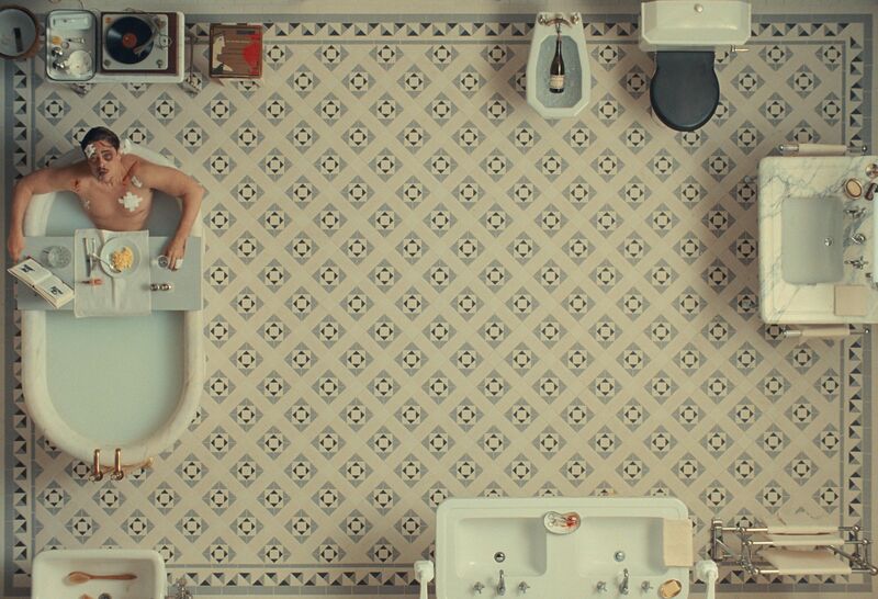 A man covered in bandages soaks in a bath in a large tiled bathroom.