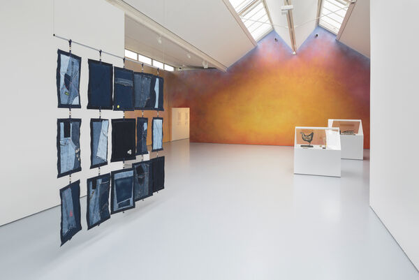 Installation image showing a vibrantly painted wall, in orange and fiery tones, at the end of gallery two. To the left an artwork consisting of sewn patchwork denim pieces hang in a grid is suspended from the ceiling. To the back right we can see two vitrines with apexes. 