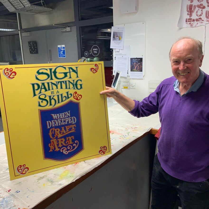 Photo of Brian Robertson in DCA Print Studio holding a sign that reads'Sign painting is a skill, when developed cratft art'
