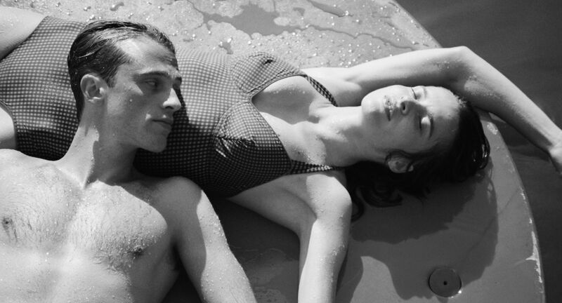 A black and white image of a man and woman laying down in swimwear, he has his head resting on her stomach and is gazing at her face, here eyes are closed and the scene feels calm